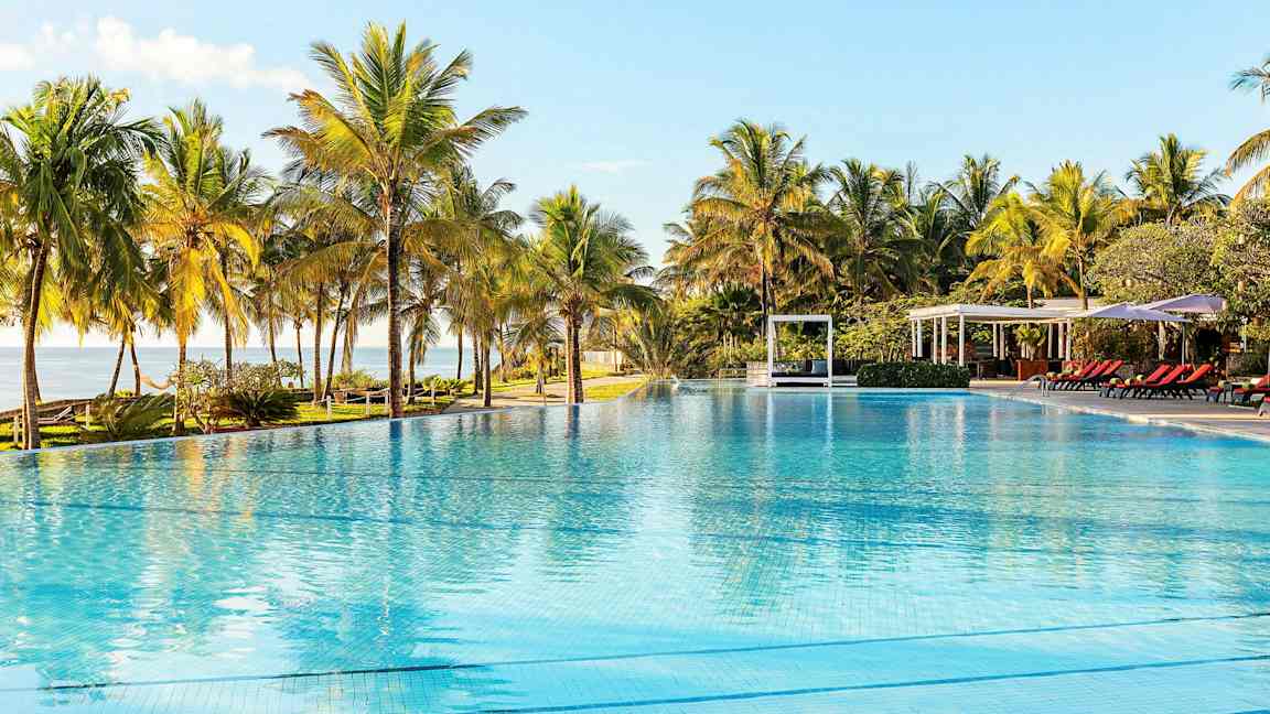 Suuri ulkouima-allas palmujen ympäröimänä, taustalla aurinkotuoleja ja paviljonkeja. (Tekoälyn luoma teksti)