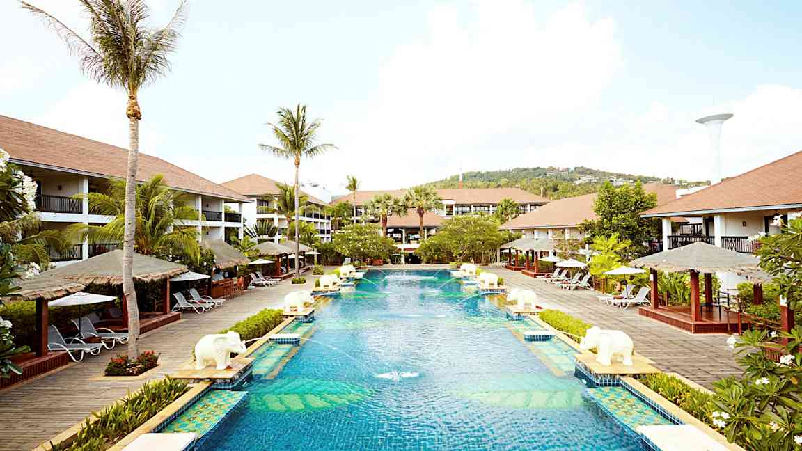 Iso uima-allas lomakeskusalueella, jota ympäröivät palmut, aurinkotuolit ja bungalow-tyyliset rakennukset. (Tekoälyn luoma teksti)