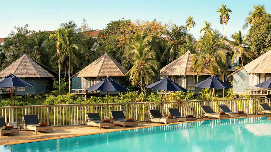 Aurinkotuoleja ja varjoja uima-altaan äärellä, jossa taustalla bungalow-tyylisiä rakennuksia ja kasvillisuutta. (Tekoälyn luoma teksti)