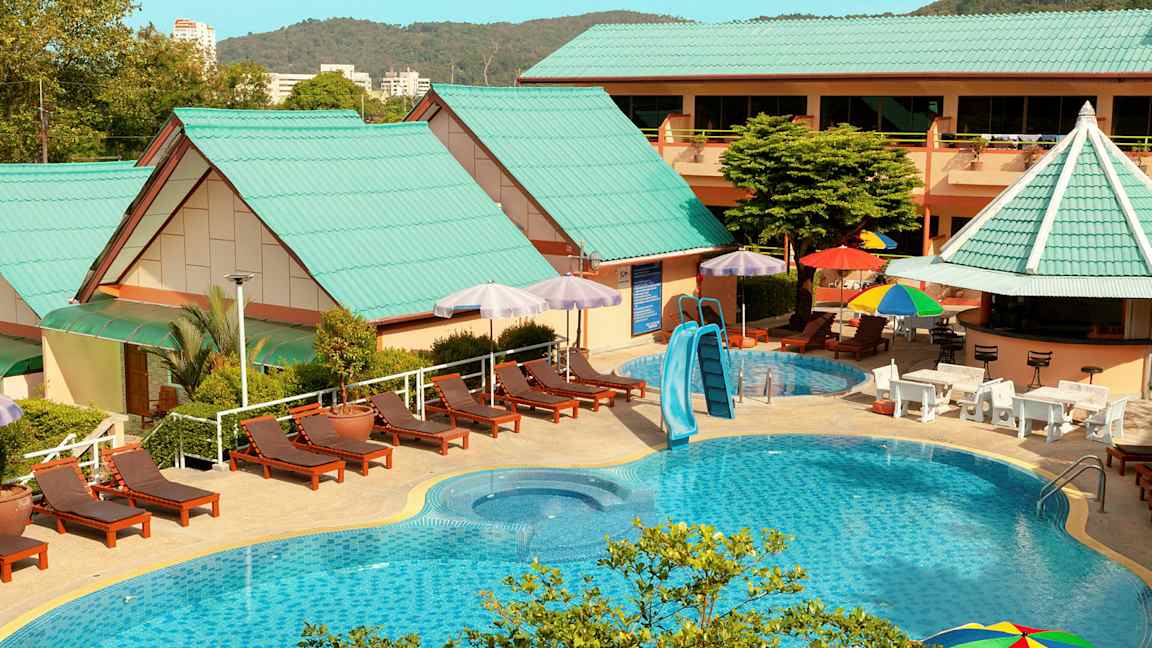 Ulkouima-allasalue aurinkotuoleilla, aurinkovarjoilla ja rakennuksilla, joissa on vihreät katot. (Tekoälyn luoma teksti)