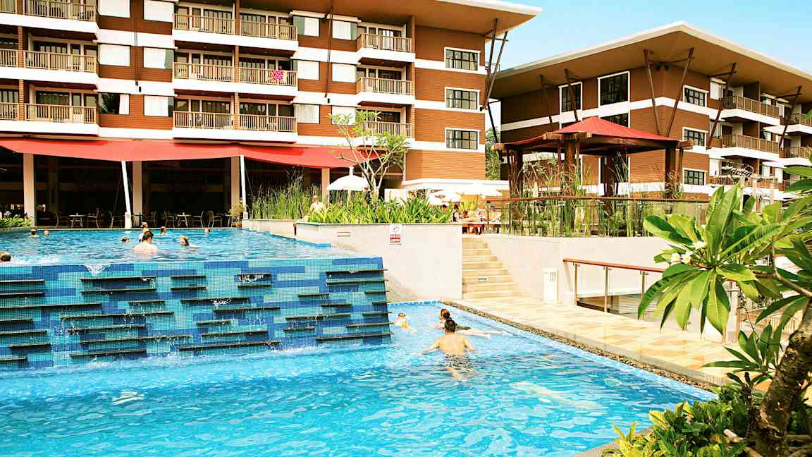 Hotelli, jossa on kaksi kerrosta, näkyy ulkouima-altaan takana, jossa ihmiset uivat. Allasalueen ympärillä on vihreitä kasveja ja terassialueita. (Tekoälyn luoma teksti)
