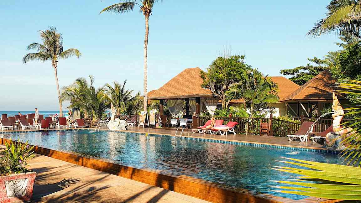 Kuva uima-allasalueesta, jossa on palmuja, aurinkotuoleja ja bungalow-tyylisiä rakennuksia taustalla. (Tekoälyn luoma teksti)