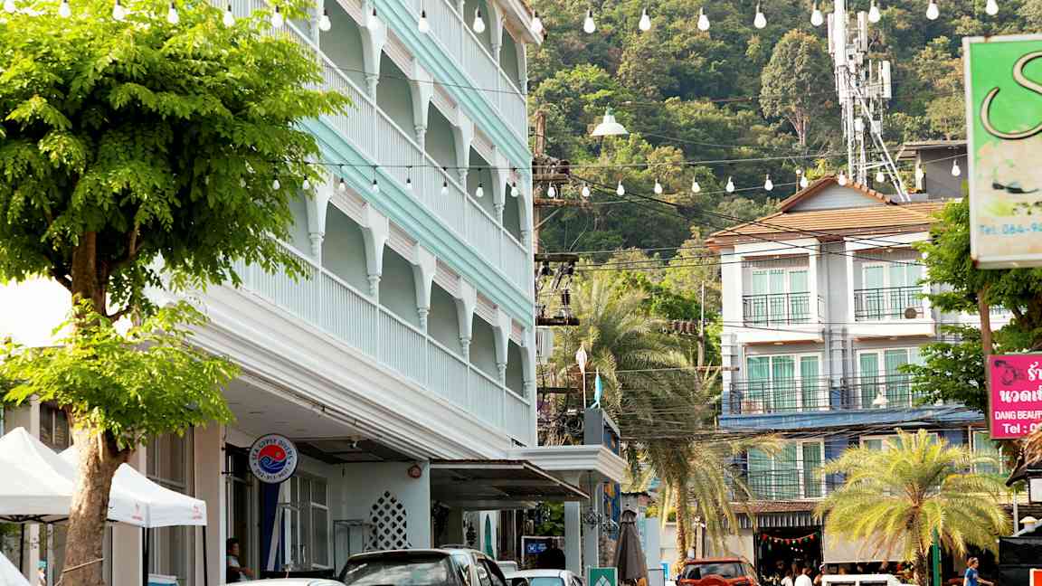 Katu, jossa on pysäköityjä autoja, rakennus sinisellä julkisivulla ja vehmaita puita. (Tekoälyn luoma teksti)