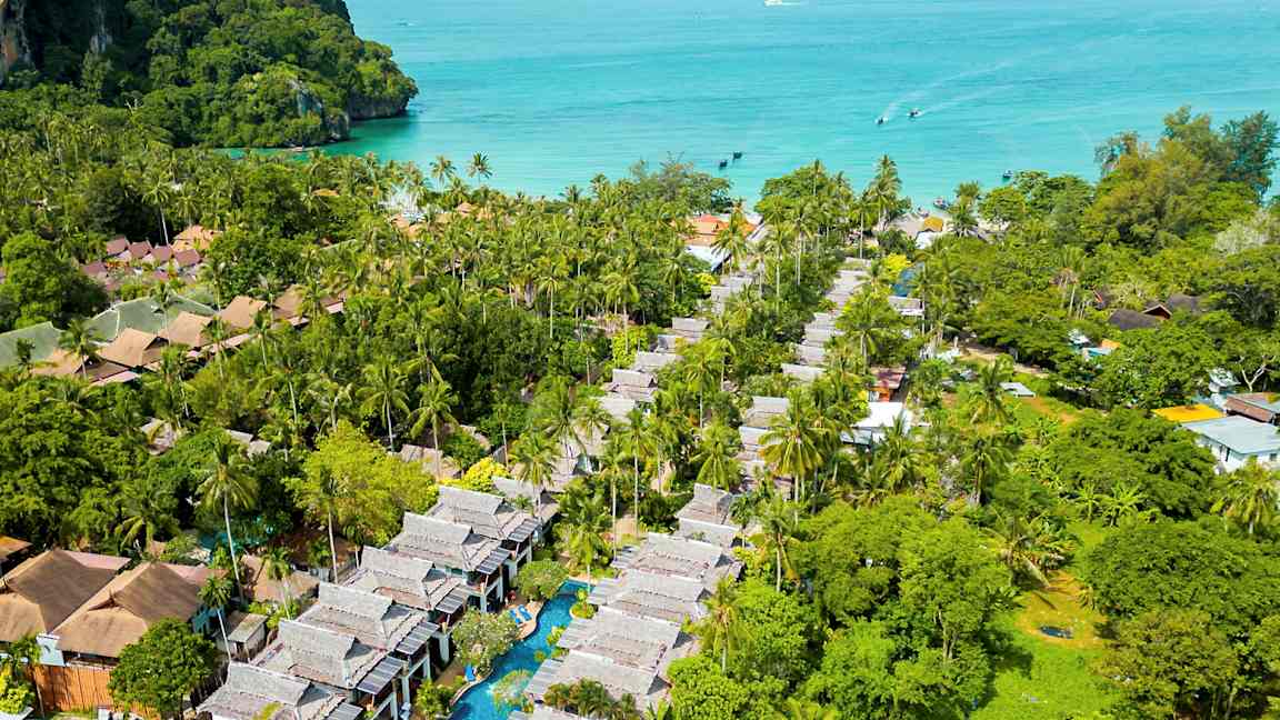 Ilmakuva trooppiselta alueelta, jossa on palmuja, rakennuksia ja uima-allas, taustalla meri. (Tekoälyn luoma teksti)