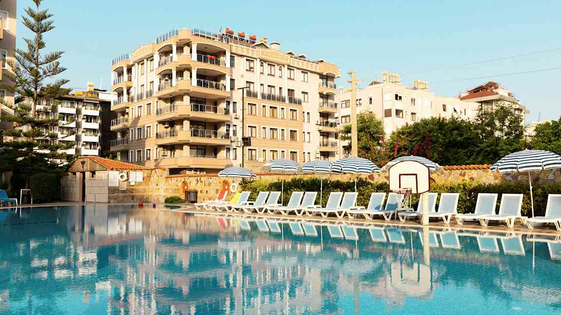 Kuva uima-altaasta, jonka sivulla aurinkotuoleja ja aurinkovarjoja, taustalla rakennus. (Tekoälyn luoma teksti)