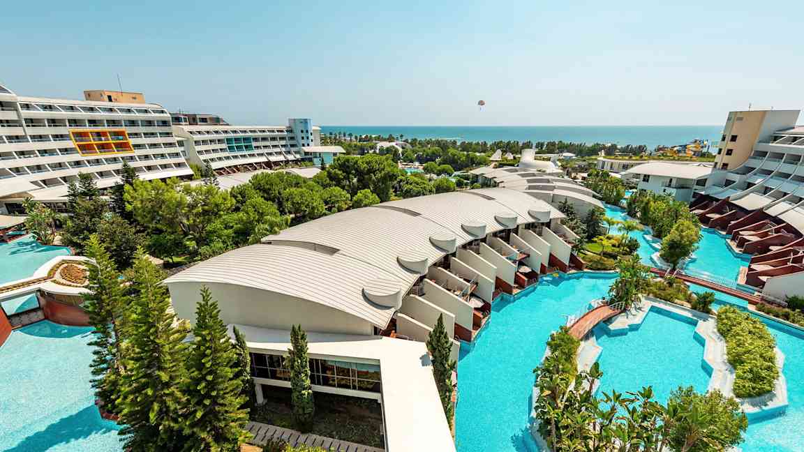 Näkymä hotellialueelta, jossa on moderneja rakennuksia, suuria uima-altaita ja vehreitä puutarhoja, jotka ulottuvat kohti merta. (Tekoälyn luoma teksti)