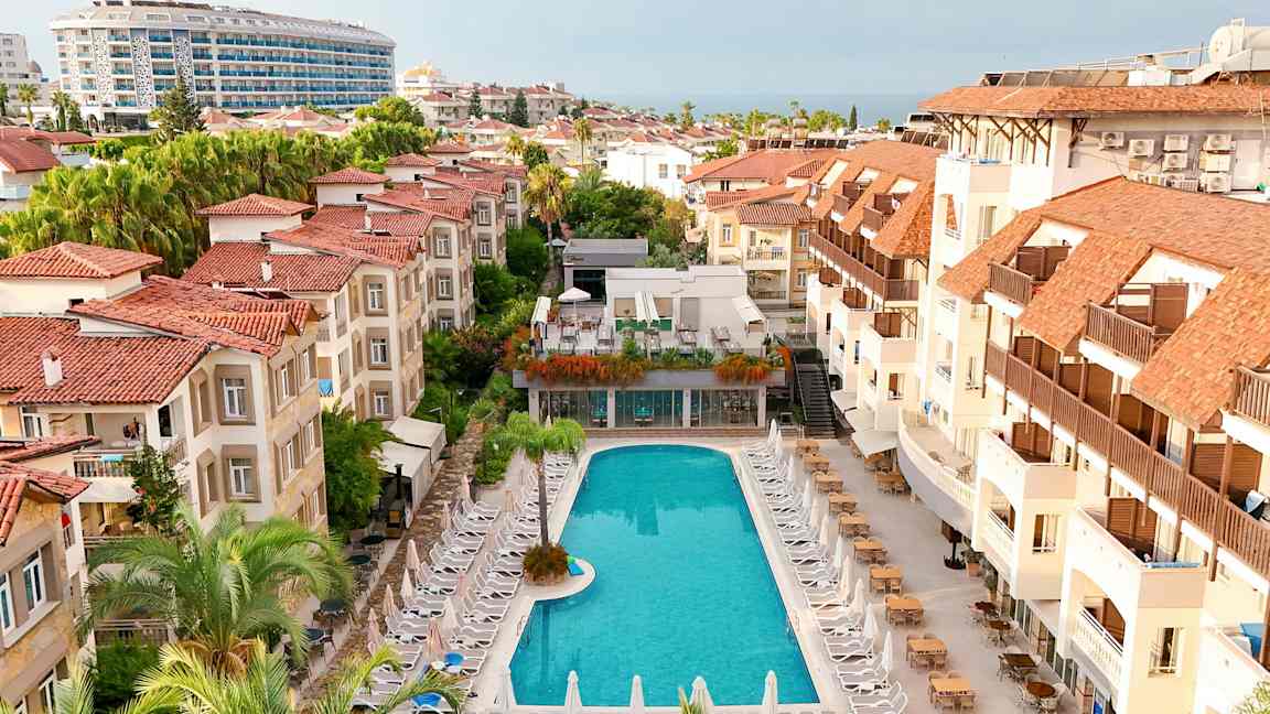 Hotellialue, jossa uima-allas ja sen ympärillä aurinkotuoleja sekä punakattoisia rakennuksia. (Tekoälyn luoma teksti)