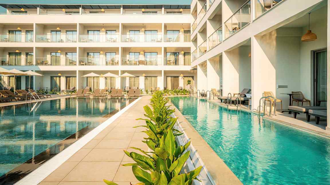 Kuva hotellista, jonka edessä on uima-allas, aurinkotuoleja ja vihreitä kasveja. (Tekoälyn luoma teksti)