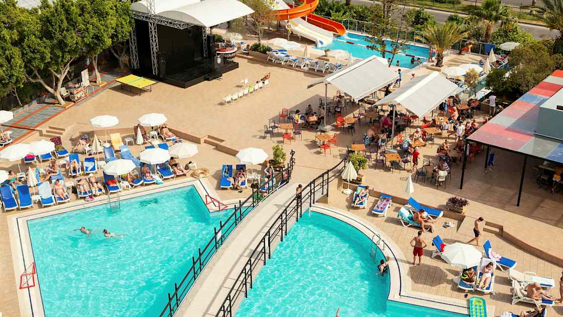 Näköala hotellialueelle, jossa uima-altaita, aurinkotuoleja ja vesiliukumäki. (Tekoälyn luoma teksti)