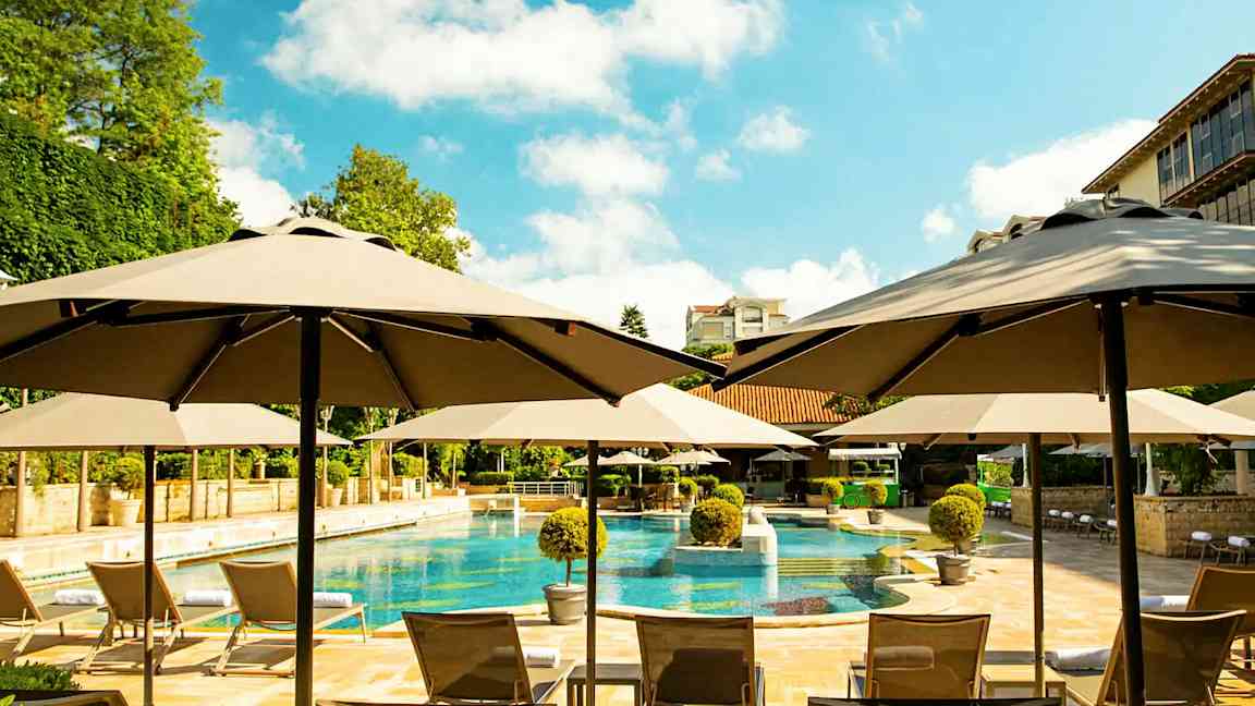 Uima-allasalue, jossa aurinkovarjoja, aurinkotuoleja ja ympäröivää vihreää. (Tekoälyn luoma teksti)