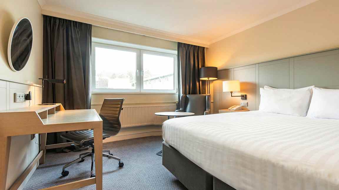 Kuva hotellihuoneesta, jossa on sänky, työpöytä ja tuoli sekä nojatuoli ikkunan vieressä. (Tekoälyn luoma teksti)