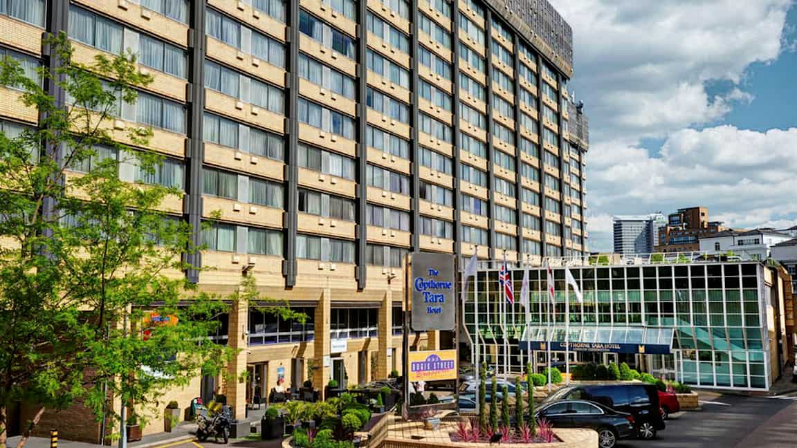 Monikerroksinen hotellirakennus, lasiovi ja pysäköintialue edessä. (Tekoälyn luoma teksti)