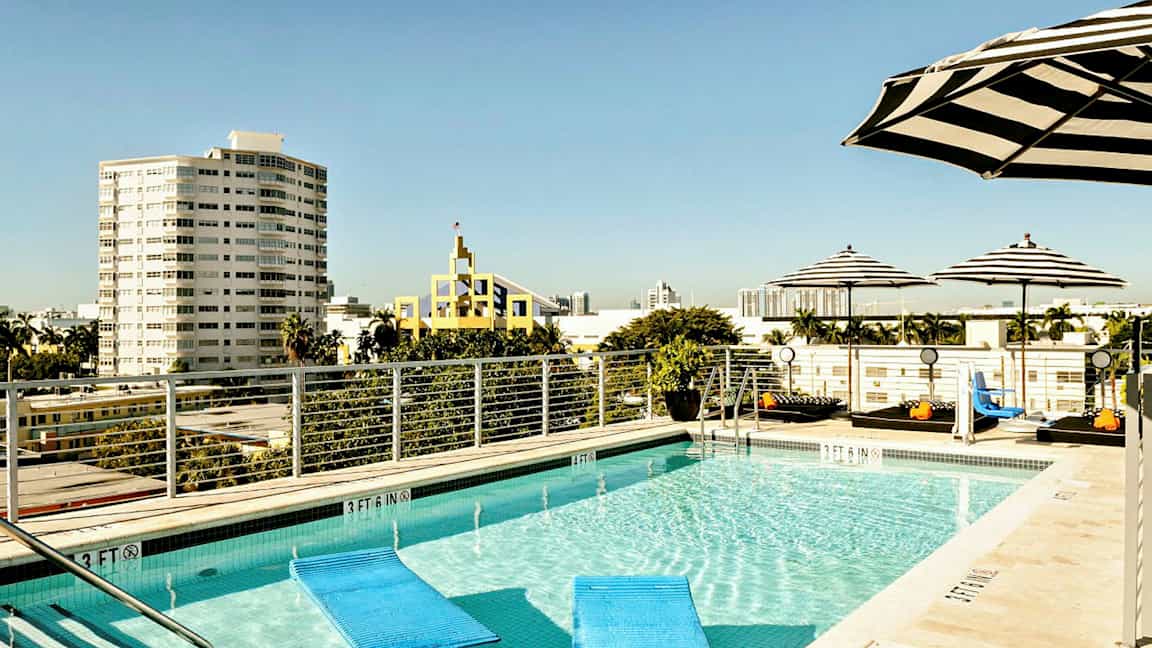 Kattouima-allas, jota ympäröivät raidalliset aurinkovarjot, taustalla kaupunkinäkymä ja useita rakennuksia. (Tekoälyn luoma teksti)