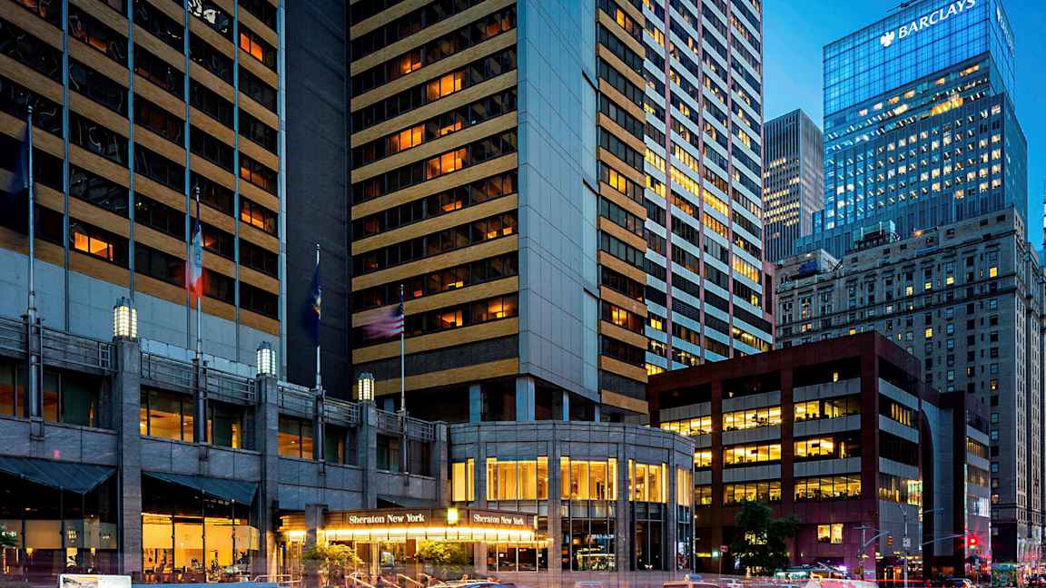 Kuva kaupunkimaisemasta, jossa on korkeita rakennuksia. Yhdessä rakennuksessa lukee "Sheraton New York" sen sisäänkäynnillä. Takseja näkyy kadulla. (Tekoälyn luoma teksti)