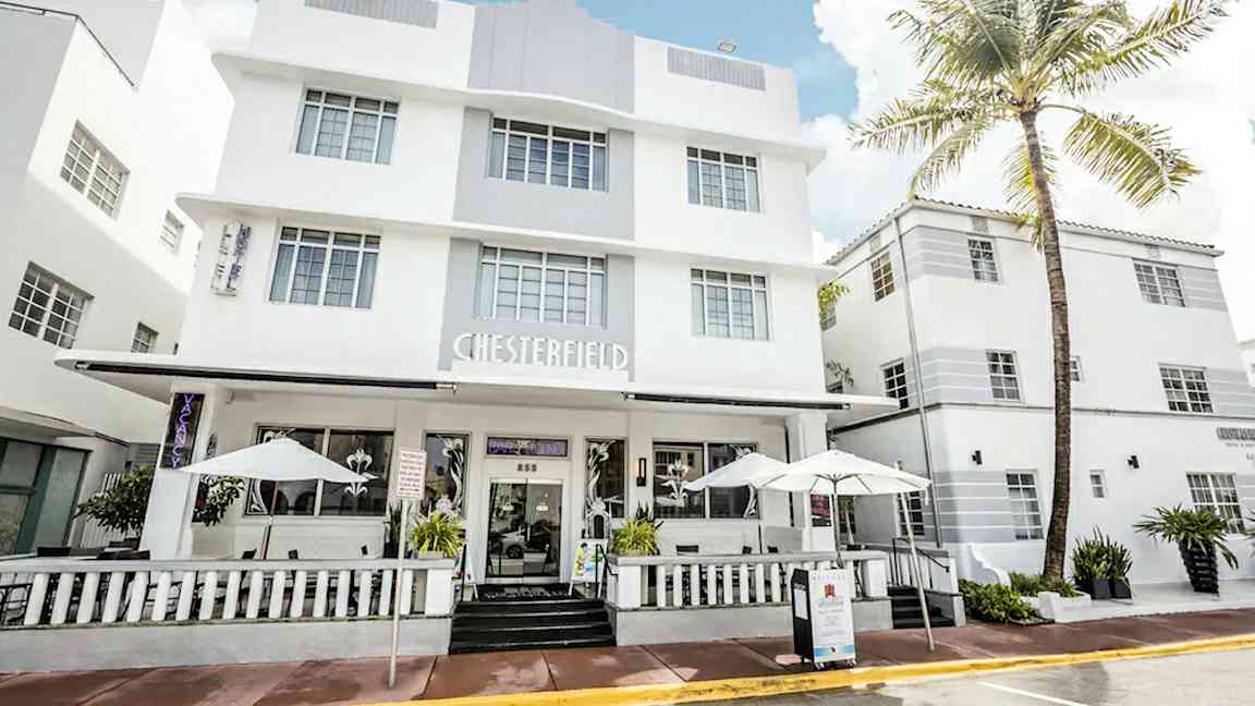 Valkoinen modernisti suunniteltu hotelli, jossa on suuret ikkunat ja kuisti aurinkovarjoilla. (Tekoälyn luoma teksti)