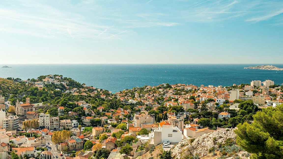 Näkymä rannikkokaupunkiin punaisine kattoineen ja vehreine alueineen, taustalla näkyy meri. (Tekoälyn luoma teksti)