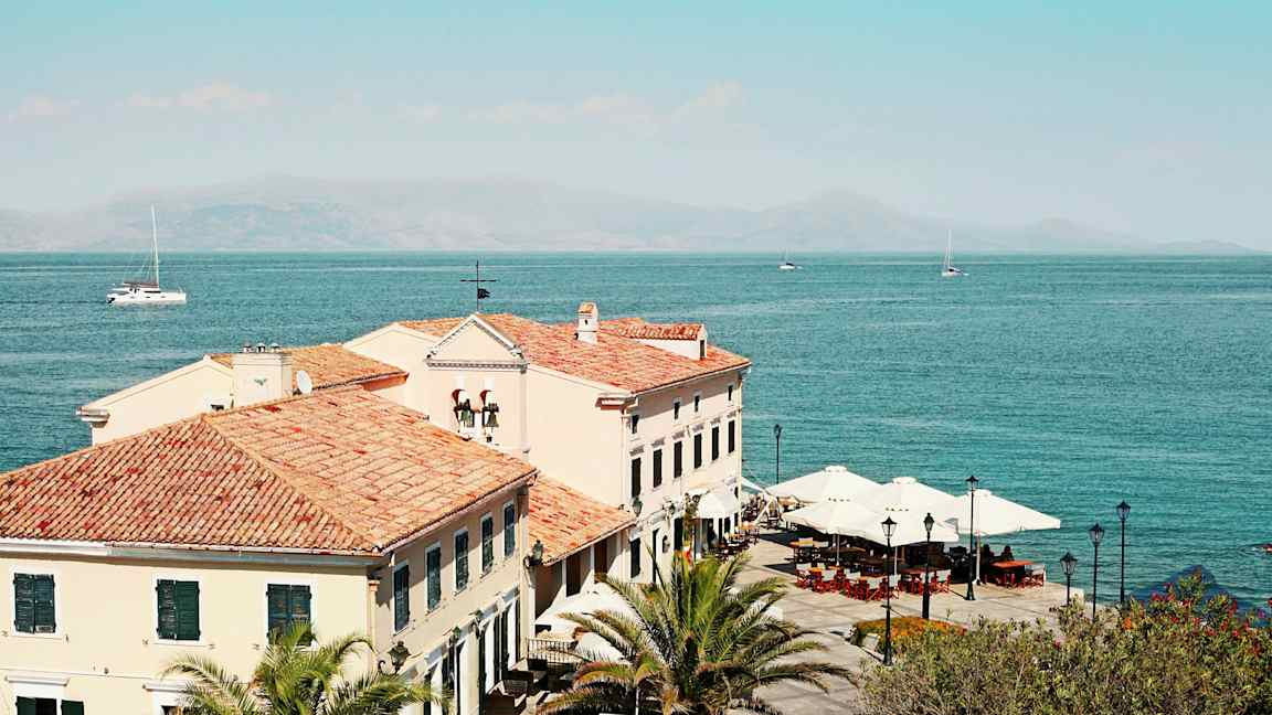 Kuva rakennuksista, joissa on tiilikatot meren rannalla, palmut ja aurinkovarjot näkyvät. (Tekoälyn luoma teksti)