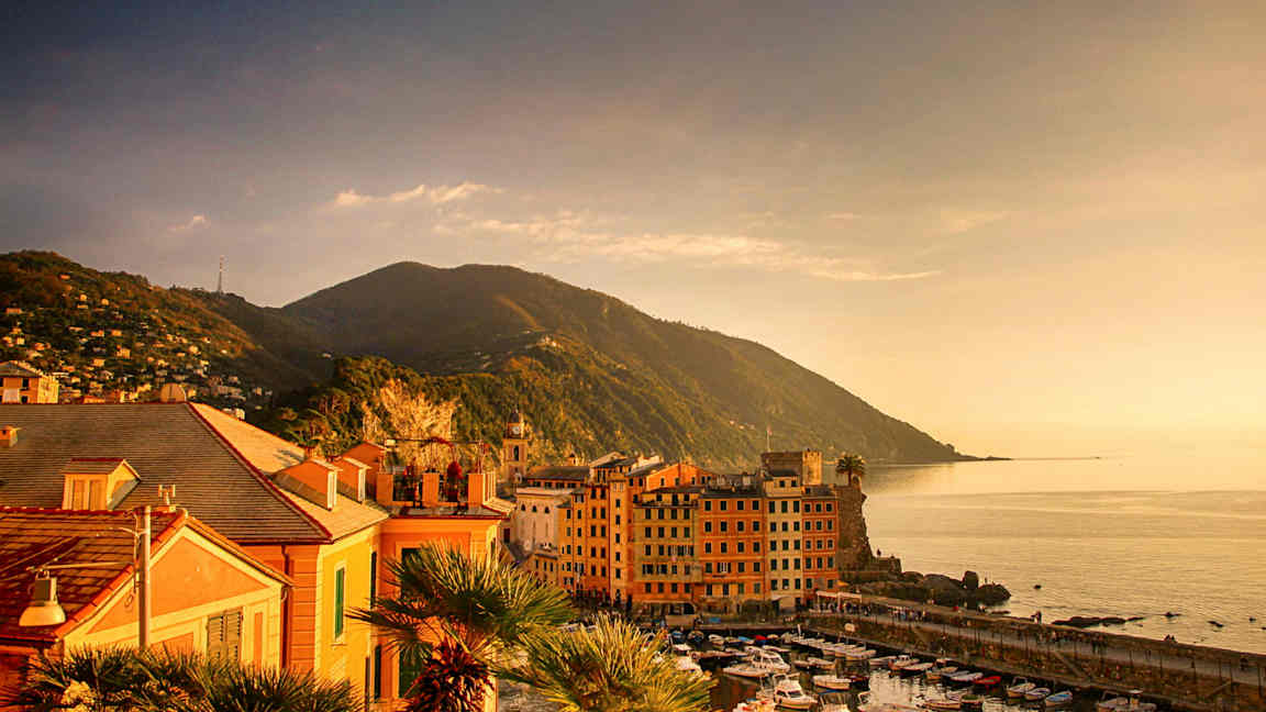 Kuva rannikkokaupungista meren äärellä, pienet veneet satamassa ja vuoria taustalla. (Tekoälyn luoma teksti)