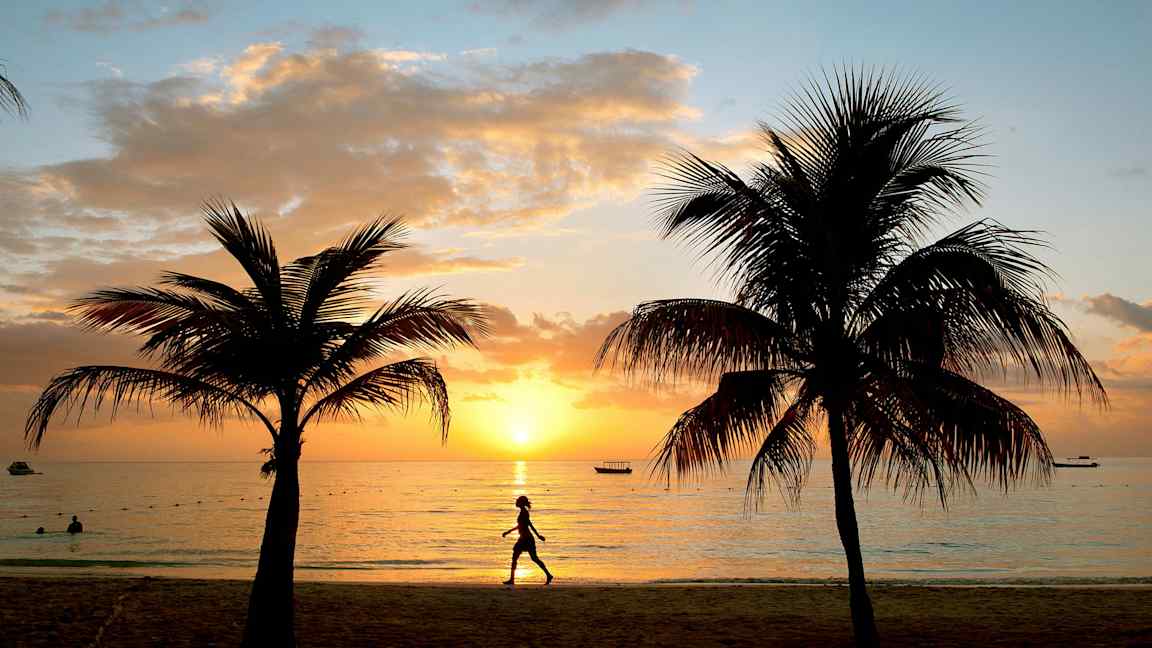 Auringonlasku rannalla palmujen keskellä ja kävelevä henkilö. (Tekoälyn luoma teksti)