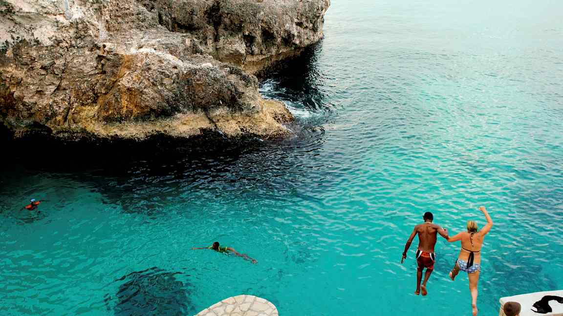 Kaksi henkilöä hyppäävät kallion reunalta siniseen veteen. (Tekoälyn luoma teksti)