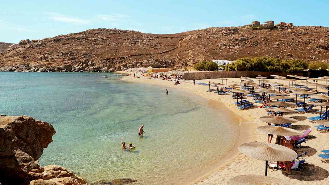 Ranta, jolla on aurinkovarjoja ja aurinkotuoleja, ihmiset uivat kirkkaassa sinisessä vedessä. (Tekoälyn luoma teksti)