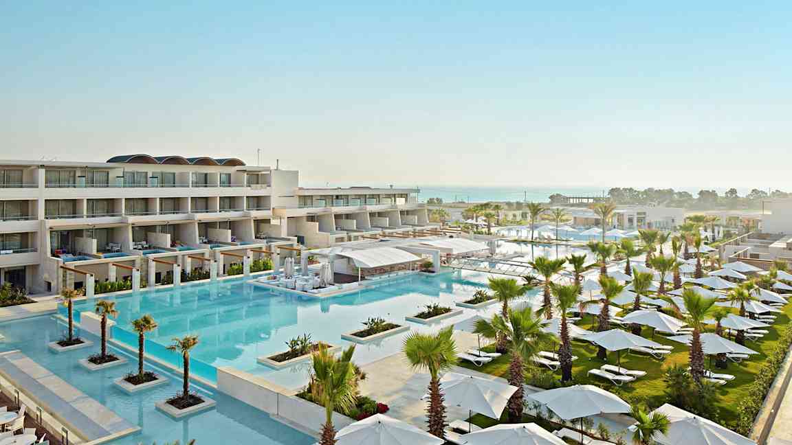 Näköala moderniin lomakeskukseen, jossa on suuri uima-allas, aurinkotuoleja, varjoja ja rakennuksia. (Tekoälyn luoma teksti)