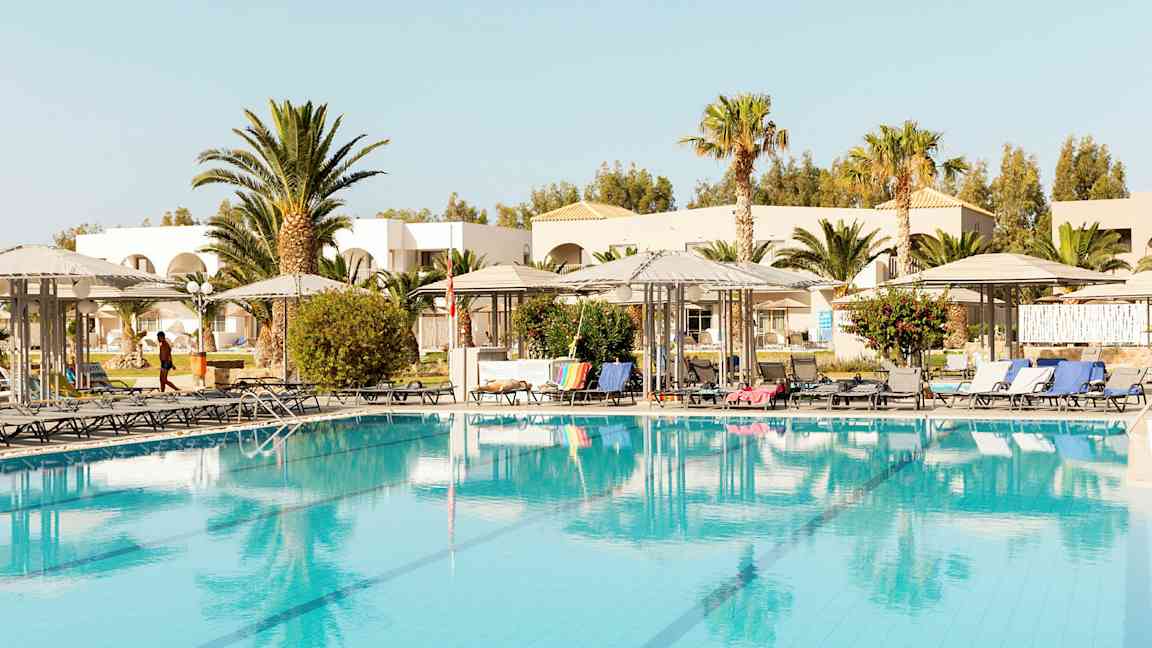 Kuva uima-altaasta aurinkotuolien ja varjojen ympäröimänä, ympärillä puita ja rakennuksia. (Tekoälyn luoma teksti)