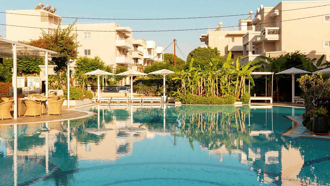 Uima-allasalue, jossa aurinkovarjoja, lepotuoleja ja rakennuksia taustalla. (Tekoälyn luoma teksti)