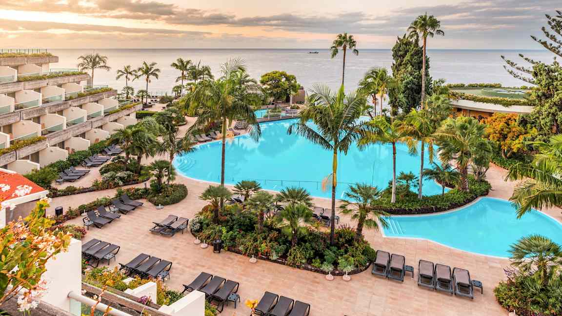 Kuva hotellikompleksista, jossa on suuri uima-allas, ympäröity aurinkotuoleilla ja trooppisilla kasveilla. Taustalla näkyy meri ja auringonlasku. (Tekoälyn luoma teksti)