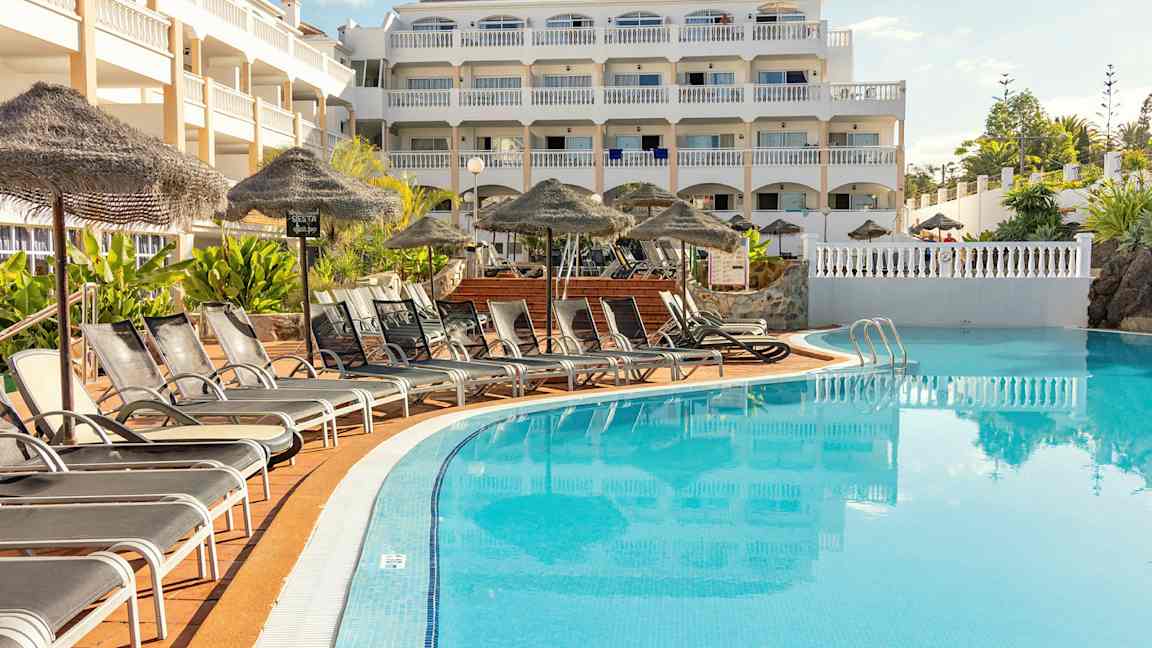 Kuvassa monikerroksinen hotellirakennus uima-altaan vieressä. Altaan edessä lepotuoleja ja aurinkovarjoja. (Tekoälyn luoma teksti)