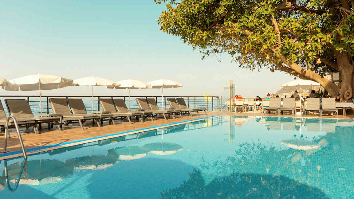 Ulkouima-allas, jonka vieressä on aurinkotuoleja ja aurinkovarjoja, sekä puu ja puuterassi istumapaikoilla. (Tekoälyn luoma teksti)