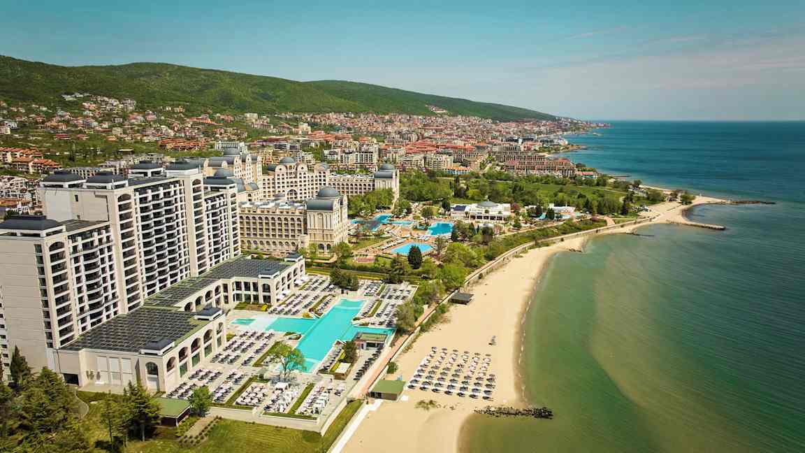 Hotellit ja uima-altaat rannalla, taustalla meri ja kaupunki. (Tekoälyn luoma teksti)