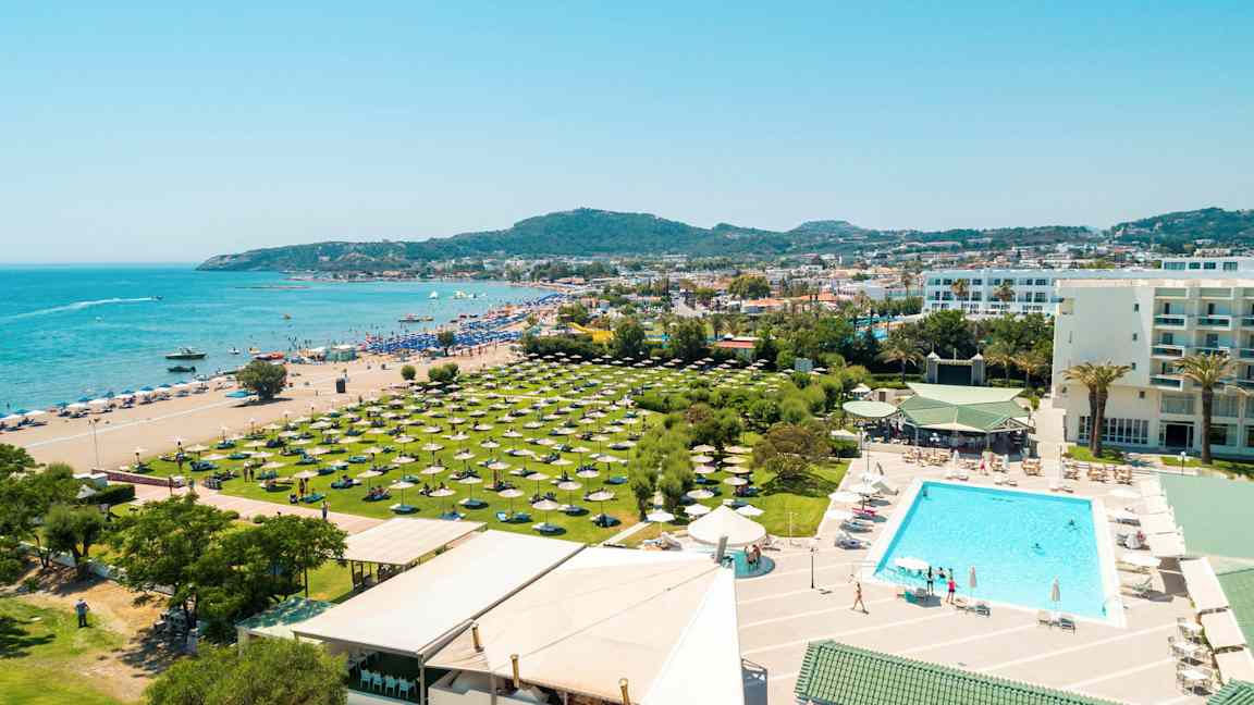 Postikortti, jossa näkyy ranta, hotellin uima-allasalue ja vihreitä alueita aurinkovarjoineen. (Tekoälyn luoma teksti)