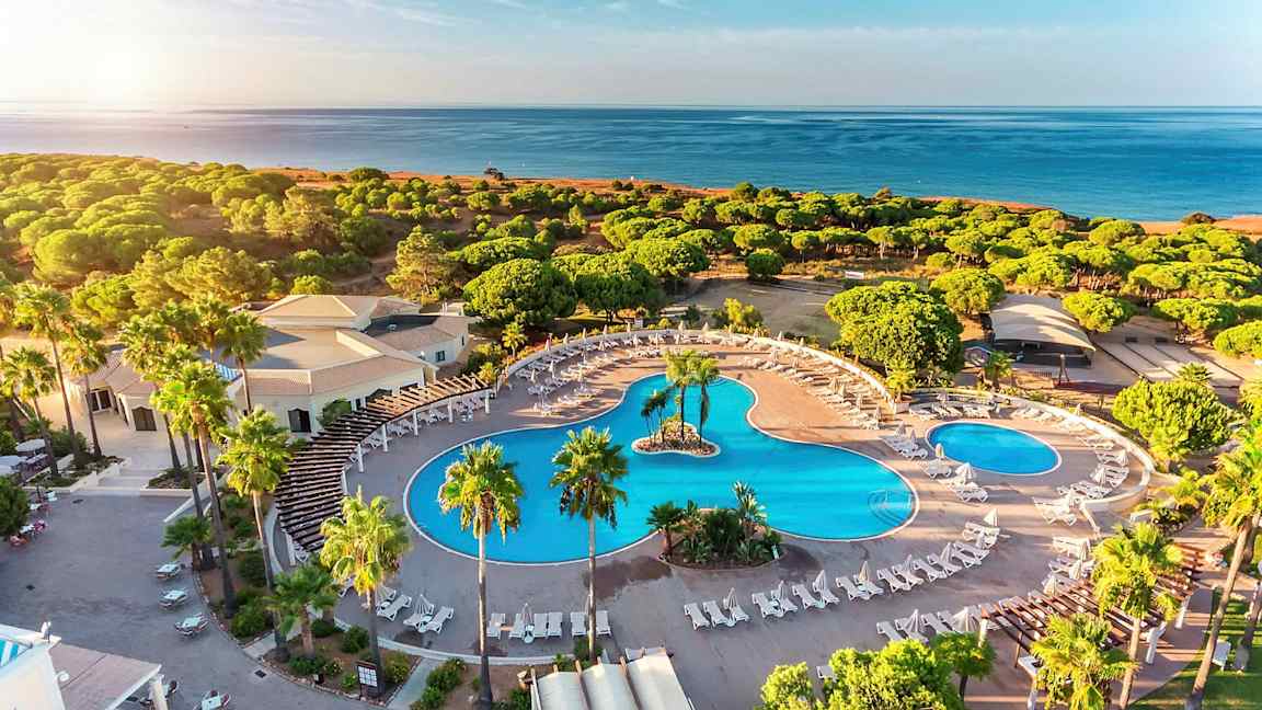 Ilmakuva hotellialueesta, jossa on suuria uima-altaita, aurinkotuoleja ja ympäröivää vehreyttä lähellä rantaa. (Tekoälyn luoma teksti)