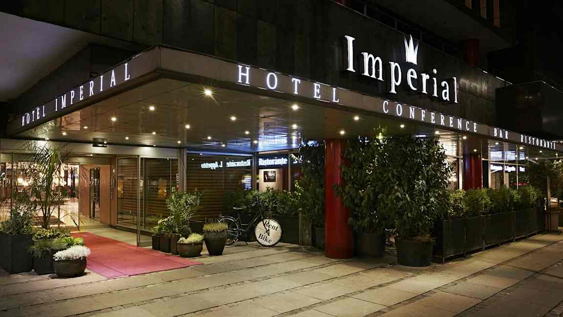Hotellin ulkokäyttö illalla kyltillä 'Hotel Imperial', ympärillä kasveja ja polkupyörä, jossa kyltti 'Rent Bike'. (Tekoälyn luoma teksti)