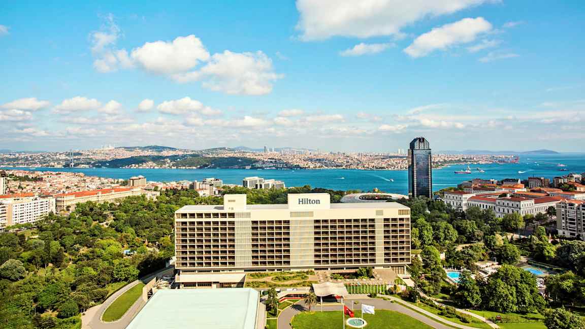 Kuvassa suuri Hilton-hotelli etualalla, jota ympäröivät viheralueet, kaupunki ja meri taustalla. (Tekoälyn luoma teksti)