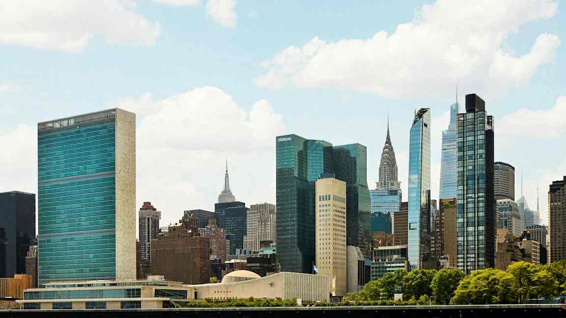 Kuvassa näkyy kaupungin siluetti, jossa on useita korkeita rakennuksia ja pilvenpiirtäjiä vehreyden ympäröimänä kirkkaalla taivaalla. (Tekoälyn luoma teksti)