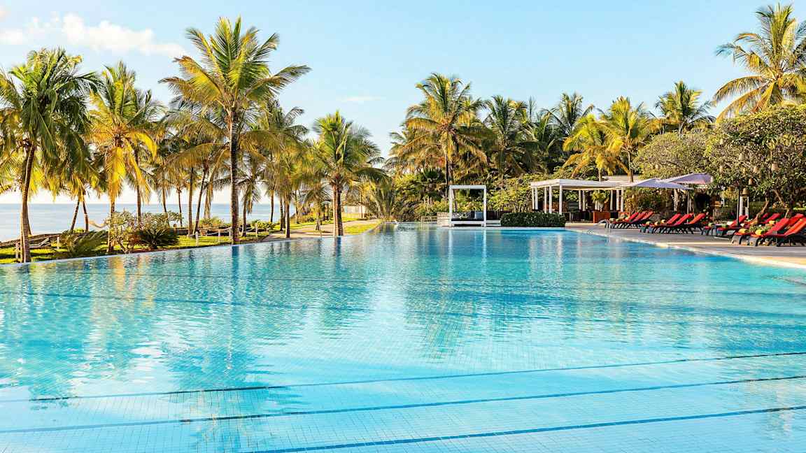 Uima-allas kirkkaalla vedellä, palmujen ja aurinkotuolien ympäröimänä, kirkkaan taivaan alla. (Tekoälyn luoma teksti)