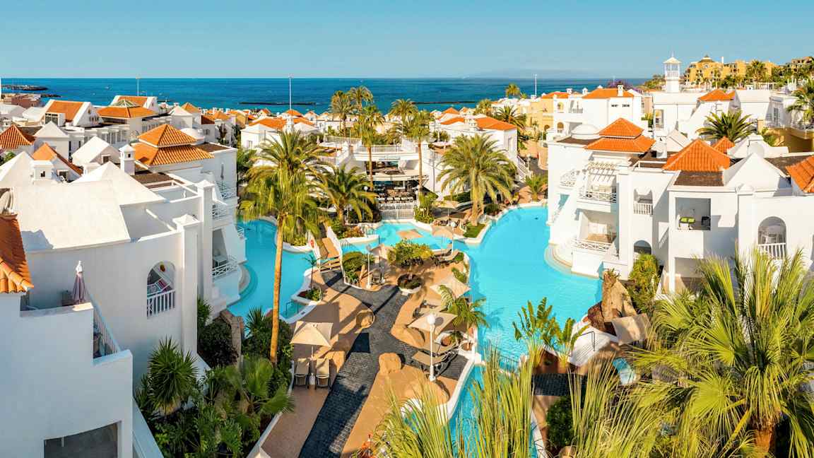 Uima-allasalue, jossa valkoiset rakennukset ja punaiset katot, ympäröity palmuilla ja näkymä merelle. (Tekoälyn luoma teksti)