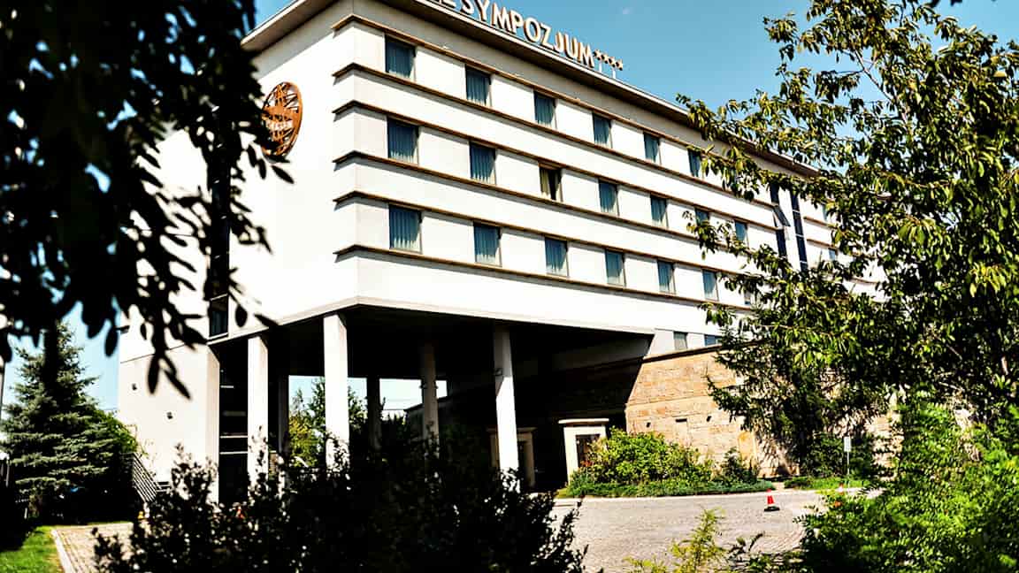 Monikerroksinen rakennus, jonka kyljessä lukee 'Hotel Sympozjum', ympäröitynä puilla ja kasvillisuudella. (Tekoälyn luoma teksti)