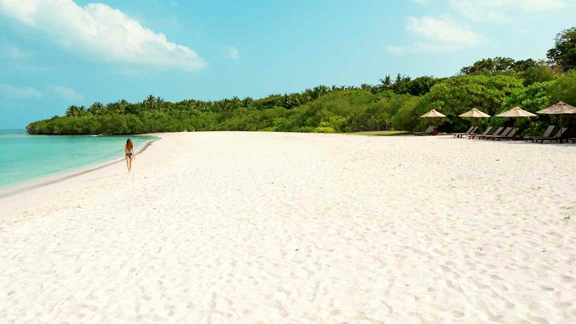 Nainen kävelee hiekkarannalla, jossa taustalla turkoosi vesi ja vihreä kasvillisuus. (Tekoälyn luoma teksti)