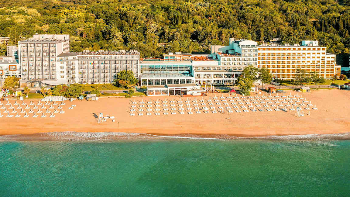 Hotelli rannalla, jossa on aurinkotuoleja ja aurinkovarjoja metsän edessä. (Tekoälyn luoma teksti)