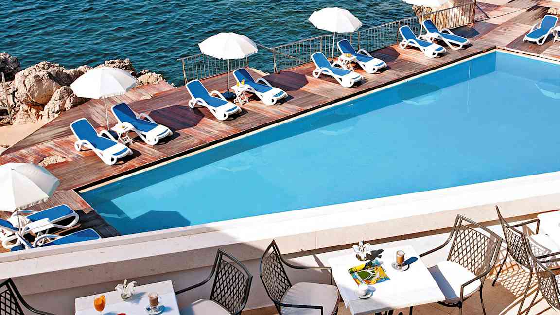 Uima-allas, jossa aurinkotuoleja ja päivänvarjoja puuterassilla meren äärellä. Etualalla näkyy pöytä tuoleineen ja juomineen. (Tekoälyn luoma teksti)