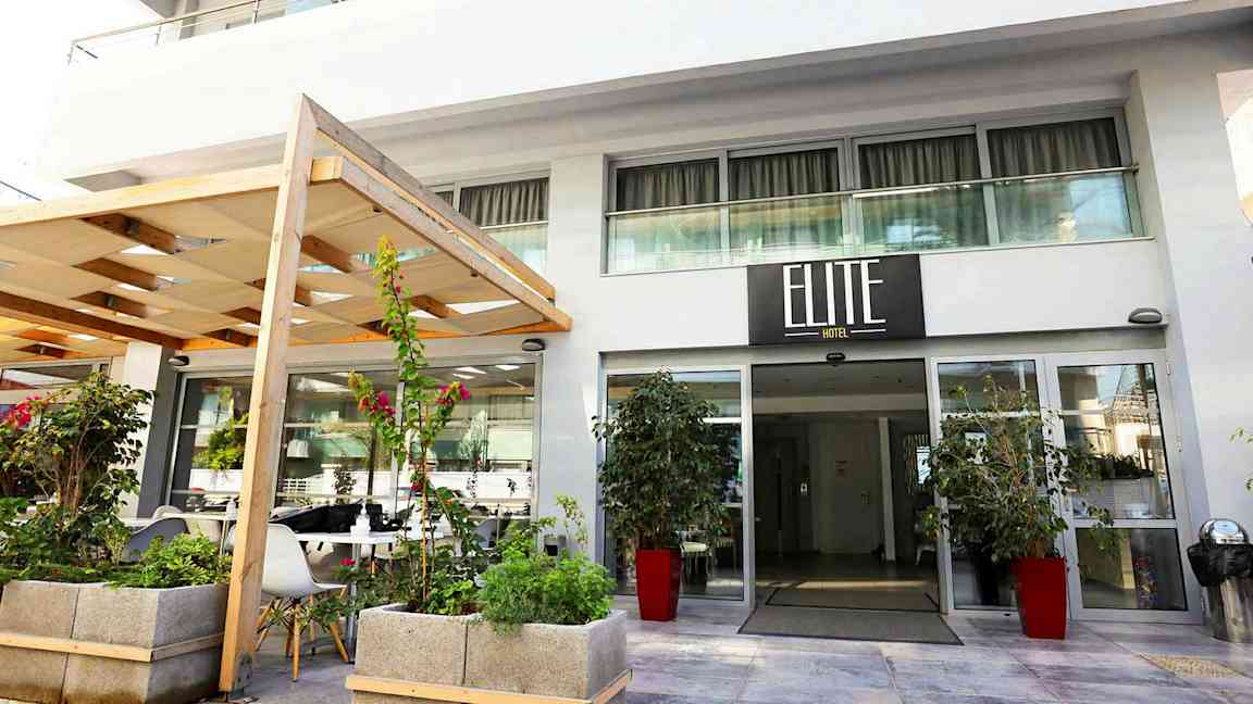 Hotellin sisäänkäynti, jossa kyltti 'Elite Hotel', ympäröitynä kasveilla ja kukilla. (Tekoälyn luoma teksti)