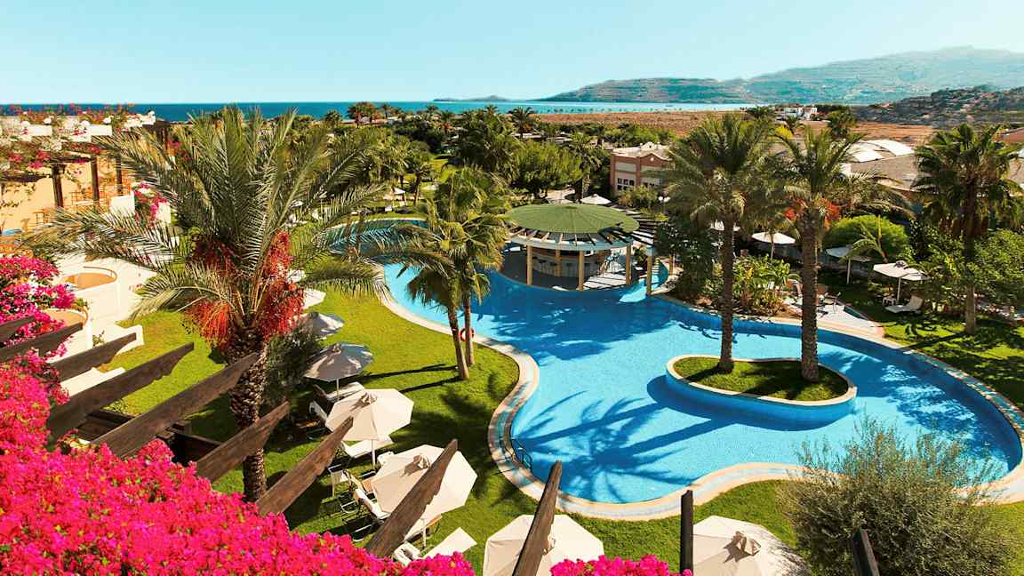 Kuva hotellialueesta, jossa on suuri uima-allas ja palmuja, sekä näkymä merelle. (Tekoälyn luoma teksti)