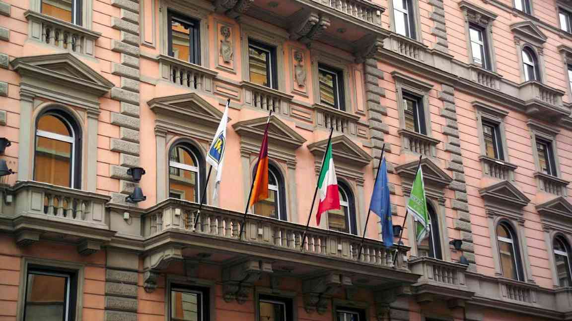 Rakennuksen julkisivu vaaleanpunaisilla ja beigeillä yksityiskohdilla, koristeltu useilla ikkunoilla ja lipuilla. (Tekoälyn luoma teksti)