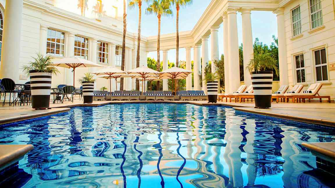 Uima-allasalue, jossa kirkasta sinistä vettä, aurinkotuoleja ja päivänvarjoja pylväiden ja palmujen ympäröimänä. (Tekoälyn luoma teksti)