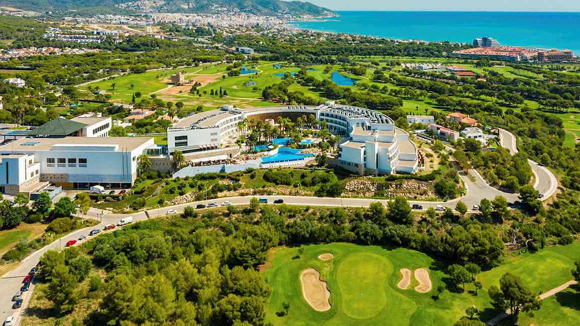 Ilmakuva hotellialueesta, jossa on uima-altaita, ympäröiviä golfkenttiä ja näköala kaupungin suuntaan rannikolle. (Tekoälyn luoma teksti)