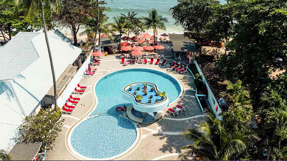 Ilmakuva uima-altaasta lomakeskuksessa, jossa on aurinkotuoleja ja varjoja. Ympärillä on trooppisia puita ja lähellä merta. (Tekoälyn luoma teksti)