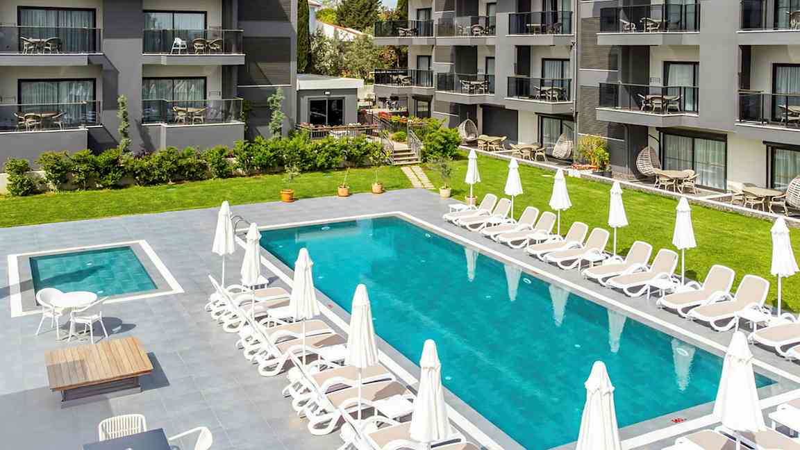 Hotellialue, jossa uima-allas, jota ympäröivät aurinkotuolit ja aurinkovarjot, taustalla rakennuksia parvekkeineen. (Tekoälyn luoma teksti)